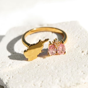 Op de afbeelding: Een gouden ring met een uitsnijding van de omtrek van Afghanistan en twee roze en heldere kristallen.
