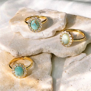 May include: Three gold rings with oval turquoise stones set in a halo of clear cubic zirconia. The rings are displayed on white rocks.