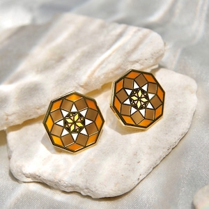 Puede incluir: Un par de pendientes de color dorado con un diseño geométrico. Los pendientes son de forma octogonal y presentan un patrón multicolor de marrón, naranja, blanco y amarillo.
