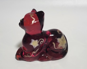 Peut inclure: Figurine de chat en verre rouge rubis en position de repos. Le chat a des décorations dorées et une tête noire. La figurine est un objet décoratif, probablement pour être exposée.