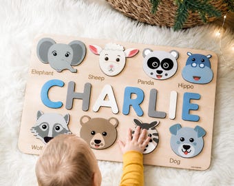 Op de afbeelding: Een rechthoekige houten naampuzzel met de naam CHARLIE in grote blauwe, grijze en witte letters. Het bord bevat acht kleurrijke uitsparingen van dierenkoppen met de labels Elephant, Sheep, Panda, Hippo, Wolf en Dog, plus een beer en zebra.