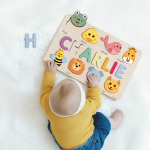 A baby is playing with a wooden puzzle featuring animal faces and the name "CHARLIE" written on it.