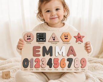 May include: A wooden educational puzzle board featuring the name EMMA in large letters, a row of five geometric shapes with faces, and a row of numbers from zero to nine, each with distinct patterns in shades of pink, black, white, and orange.