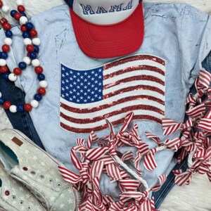 May include: A light blue t-shirt with a sequined American flag design. The shirt is distressed and has a faded look. The flag is red, white, and blue. There is a red, white, and blue beaded necklace, a red and white striped ribbon, and a blue denim jacket in the background.