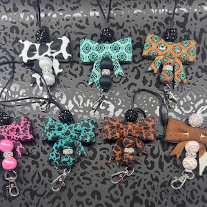 May include: A collection of seven lanyards featuring decorative bows in various patterns including cow print, geometric teal, orange tribal, pink leopard, teal leopard, brown leopard, and brown fur, each accented with beads and a metal clasp.
