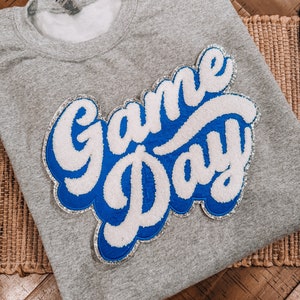 May include: A gray sweatshirt with a blue and white felt applique that says "Game Day" in a retro font.