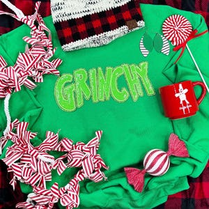 May include: Green sweatshirt with the word "Grinchy" in sparkly green letters. The sweatshirt is surrounded by red and white striped fabric, a red and white candy cane, a red mug with a white jingle bell logo, a white and black knit hat, and red and white earrings.