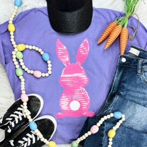 May include: Purple t-shirt with a pink sequin bunny graphic. The shirt is paired with jeans, a black baseball cap, and a beaded necklace with pastel colored beads.