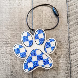 May include: A white paw print ornament with a blue and white checkered pattern. The ornament has a black string attached to it.