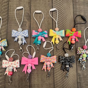 May include: A collection of colorful bow-shaped keychains with various patterns and bead accents. Each keychain features a different design, including floral, polka dot, and animal print bows, with a lobster clasp for easy attachment.