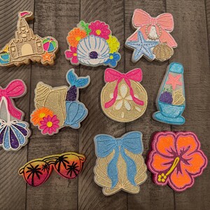 May include: A collection of colorful, glittery beach-themed embellishments. Designs include a sandcastle, seashells, a starfish, a mermaid tail, sunglasses, a lava lamp, and a hibiscus flower. The embellishments are in shades of pink, blue, orange, yellow, and gold.