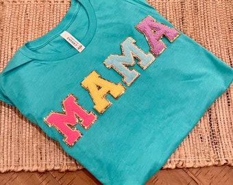 May include: A turquoise blue t-shirt with the word "MAMA" in large, colorful, fuzzy letters. The letters are outlined in gold glitter.