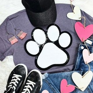 May include: A purple t-shirt with a white paw print applique. The shirt is paired with a black baseball cap, blue jeans, and a string of pink and white heart-shaped decorations.