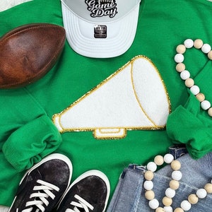 May include: Green sweatshirt with a white megaphone with gold glitter trim. The sweatshirt has the text "Game Day" on a white baseball cap. There are also wooden beads in the image.