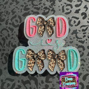May include: A decorative glittery sign with the words "Good is Good" in pink and teal. The letters are outlined with glitter and feature leopard print bows. A small heart is between the words.
