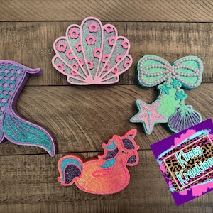 May include: A collection of glittery mermaid and sea-themed decorations. Includes a mermaid tail, seashell, bow, starfish, seashell, unicorn, and a sign that says "Kimes Kreations". The items are in shades of pink, teal, and purple.