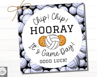 May include: A white circle with black text that says "Chip! Chip! HOORAY It's Game Day! GOOD LUCK!"  The circle is surrounded by a pattern of black and white volleyballs. There are two brown cookies with white frosting on either side of a white volleyball.