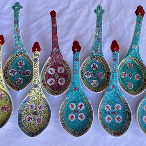 May include: A collection of eight ceramic Chinese soup spoons. Each spoon features a long handle and a rounded bowl, with intricate designs in teal, yellow, and red. The spoons are decorated with floral and geometric patterns and Chinese characters.