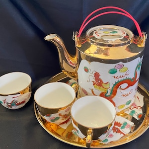May include: A gold tea set with a teapot, three cups, and a saucer. The teapot has a red handle and a dragon design. The cups and saucer also have dragon designs.