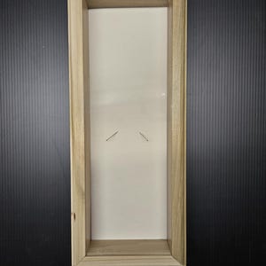 May include: A rectangular wooden shadow box with a light-colored interior and a clear front. The frame is made of light-colored wood. Two small pins are visible inside the box. The shadow box is empty and ready for display.