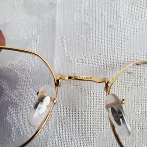 Pu&ograve; includere: Primo piano di occhiali con montatura dorata e lenti trasparenti. Il ponte degli occhiali riporta la scritta "RAY BAN 49 20". I naselli sono trasparenti e le aste sono sottili e dorate.