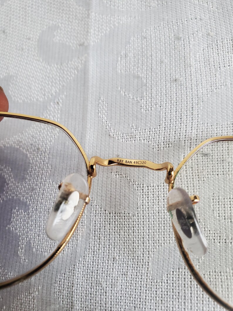 Pu&ograve; includere: Primo piano di occhiali con montatura dorata e lenti trasparenti. Il ponte degli occhiali riporta la scritta "RAY BAN 49 20". I naselli sono trasparenti e le aste sono sottili e dorate.