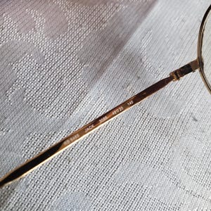 Pu&ograve; includere: Primo piano di occhiali dorati con lenti rotonde. L'asta mostra il testo "RB 6465 JACK 3086 49 20 140". Gli occhiali hanno una montatura metallica sottile, su uno sfondo chiaro e strutturato.