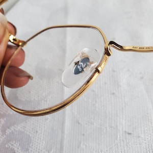 Pu&ograve; includere: Primo piano di occhiali con montatura dorata e lenti trasparenti. Il logo Ray-Ban &egrave; visibile sui naselli. La montatura &egrave; di un caldo colore oro. Il testo "RAY BAN 49" &egrave; stampato sull'asta.