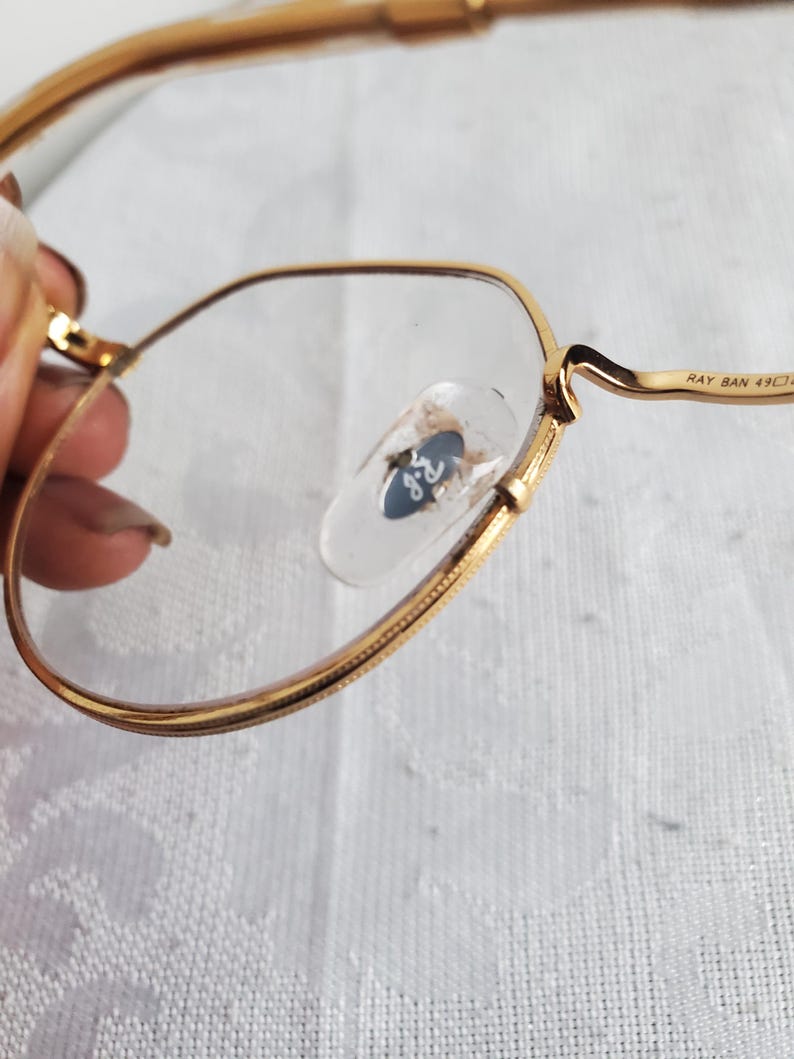 Pu&ograve; includere: Primo piano di occhiali con montatura dorata e lenti trasparenti. Il logo Ray-Ban &egrave; visibile sui naselli. La montatura &egrave; di un caldo colore oro. Il testo "RAY BAN 49" &egrave; stampato sull'asta.