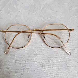 Pu&ograve; includere: Occhiali con montatura dorata di forma rotonda classica. Gli occhiali hanno una montatura metallica sottile e lenti trasparenti. Le aste sono di colore chiaro e si estendono fino alle orecchie. Gli occhiali poggiano su una superficie bianca a motivi.