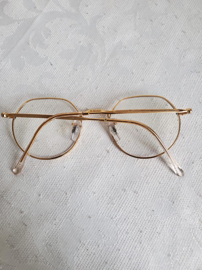 Pu&ograve; includere: Occhiali con montatura dorata di forma rotonda classica. Gli occhiali hanno una montatura metallica sottile e lenti trasparenti. Le aste sono di colore chiaro e si estendono fino alle orecchie. Gli occhiali poggiano su una superficie bianca a motivi.