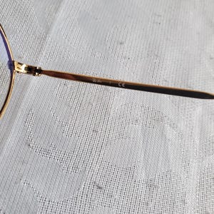 Pu&ograve; includere: Primo piano di occhiali da sole aviator dorati. La montatura &egrave; sottile e metallica, con una classica forma a goccia. Le lenti sono di colore azzurro chiaro. L'asta presenta la scritta "Ray-Ban" e "CE".