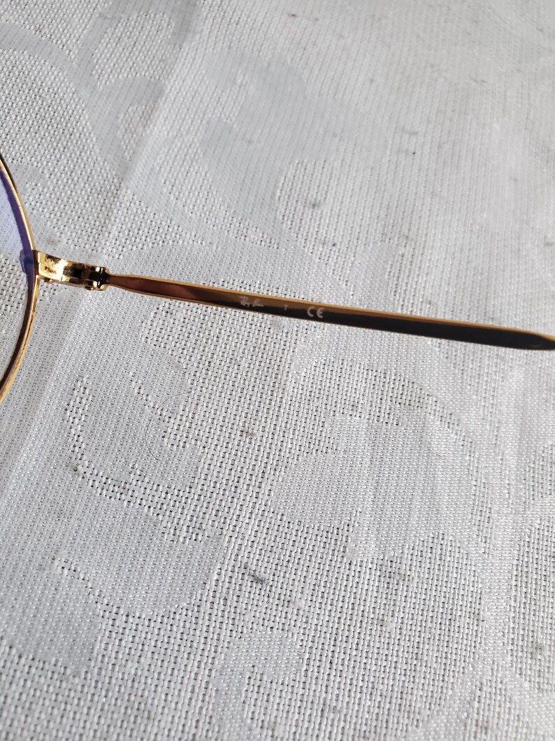 Pu&ograve; includere: Primo piano di occhiali da sole aviator dorati. La montatura &egrave; sottile e metallica, con una classica forma a goccia. Le lenti sono di colore azzurro chiaro. L'asta presenta la scritta "Ray-Ban" e "CE".