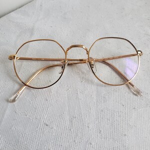 Pu&ograve; includere: Occhiali con montatura dorata con forma di lente rotonda e ottagonale. Gli occhiali hanno una sottile montatura dorata e naselli e aste trasparenti. Le lenti sono trasparenti. Gli occhiali sono appoggiati su una superficie bianca a motivi.