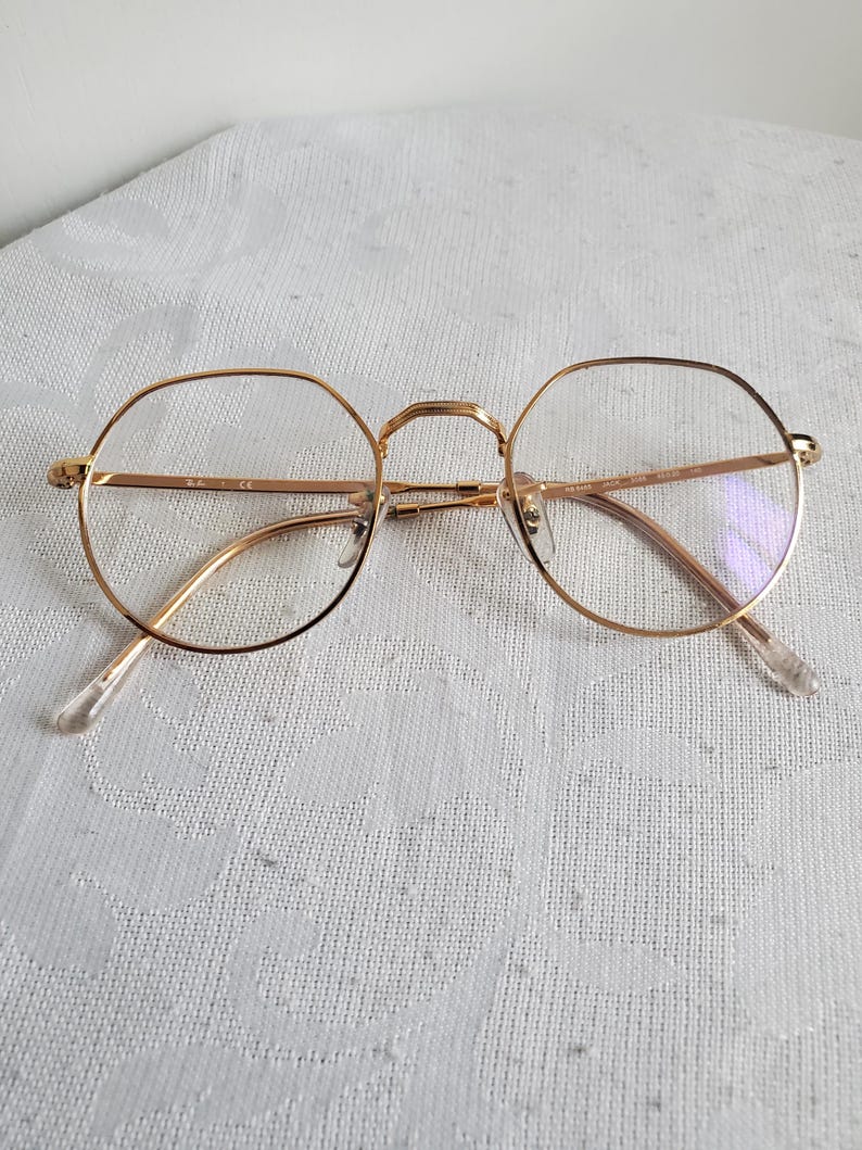 Pu&ograve; includere: Occhiali con montatura dorata con forma di lente rotonda e ottagonale. Gli occhiali hanno una sottile montatura dorata e naselli e aste trasparenti. Le lenti sono trasparenti. Gli occhiali sono appoggiati su una superficie bianca a motivi.