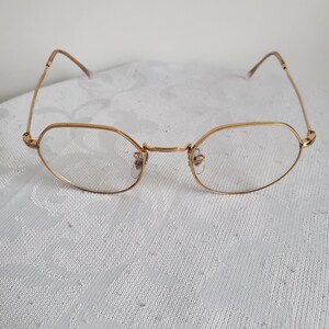 Pu&ograve; includere: Occhiali con montatura dorata e lenti ovali. Le montature sono di un caldo color oro e le aste sono sottili e delicate. Gli occhiali sono appoggiati su uno sfondo bianco strutturato. Lo stile generale &egrave; vintage e classico.