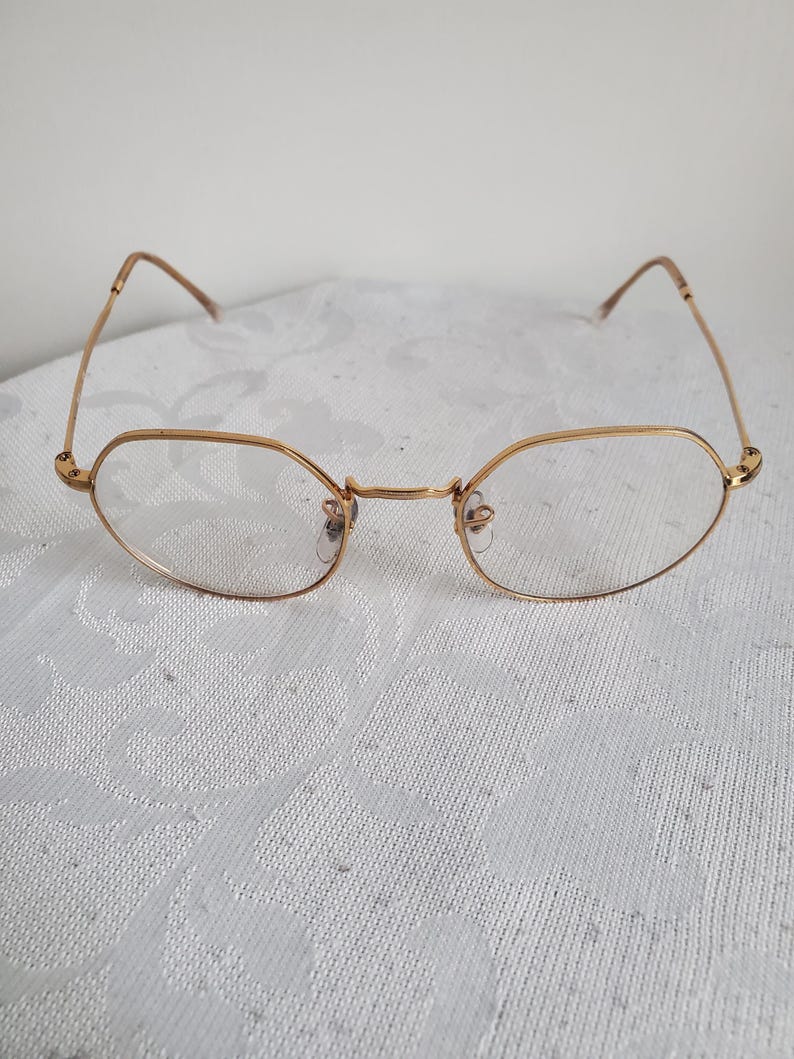 Pu&ograve; includere: Occhiali con montatura dorata e lenti ovali. Le montature sono di un caldo color oro e le aste sono sottili e delicate. Gli occhiali sono appoggiati su uno sfondo bianco strutturato. Lo stile generale &egrave; vintage e classico.