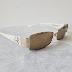 Peut inclure: Lunettes de soleil à monture blanche avec des détails dorés et des verres bruns. Les branches sont blanches avec un emblème doré. Les verres sont rectangulaires et la monture est dorée. Sur une surface blanche à motifs.