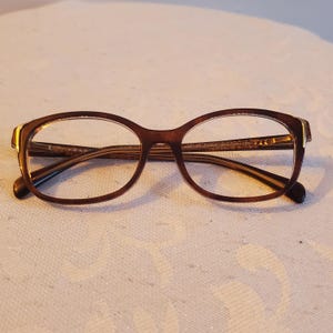 Puede incluir: Gafas ovaladas marrones con un acento dorado en las patillas. Las monturas son de color marrón oscuro y las lentes son transparentes. Las gafas están sobre una superficie blanca.