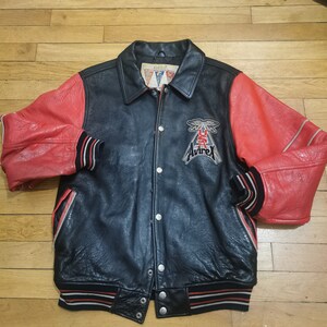 May include: A black leather varsity jacket with red leather sleeves. The jacket has a white and red striped rib knit collar, cuffs, and hem. The jacket has a snap closure and features a large embroidered patch on the left chest that says "Avirex" with a winged eagle and "USA" underneath.