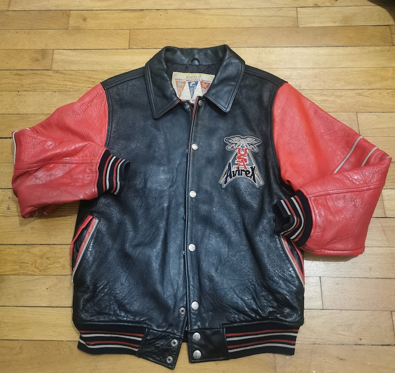 May include: A black leather varsity jacket with red leather sleeves. The jacket has a white and red striped rib knit collar, cuffs, and hem. The jacket has a snap closure and features a large embroidered patch on the left chest that says "Avirex" with a winged eagle and "USA" underneath.