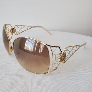 May include: Gold-tone sunglasses with gradient brown lenses. The frames feature intricate filigree and decorative floral accents. The lenses are a rounded rectangular shape. The arms are a light gold color.