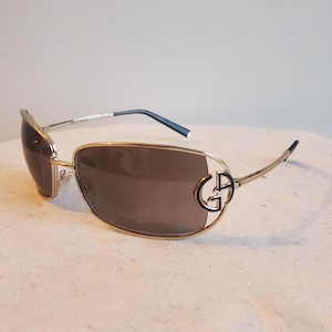 Könnte beinhalten: Goldfarbene Sonnenbrille mit braunen Gläsern. Die Fassungen haben ein markantes Design mit einem stilisierten "G"-Logo an den Bügeln. Die Bügel sind dünn und biegen sich um die Ohren. Die Gläser haben eine rechteckige Form.