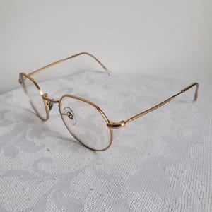 Pu&ograve; includere: Occhiali dorati con montatura ovale e aste sottili. Gli occhiali hanno lenti trasparenti e un piccolo ponte nasale. La montatura sembra essere in metallo. Gli occhiali sono appoggiati su una superficie bianca a motivi.