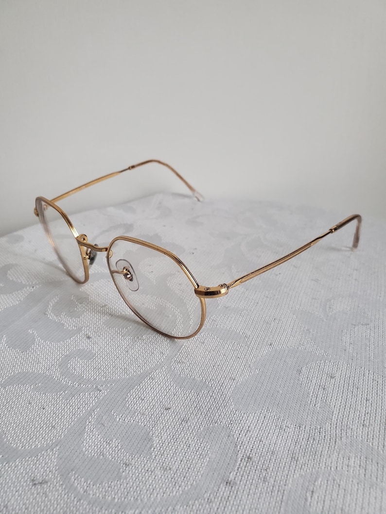 Pu&ograve; includere: Occhiali dorati con montatura ovale e aste sottili. Gli occhiali hanno lenti trasparenti e un piccolo ponte nasale. La montatura sembra essere in metallo. Gli occhiali sono appoggiati su una superficie bianca a motivi.