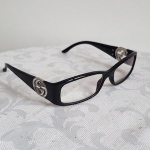 May include: Black eyeglasses with a rectangular frame. The temples feature a silver-colored logo. The glasses are set against a white background.