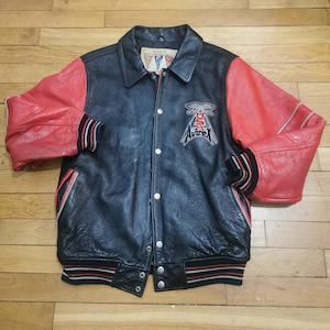 May include: A black leather varsity jacket with red leather sleeves. The jacket has a snap closure, two pockets, and a ribbed collar, cuffs, and hem. The jacket has a large "AVIREX" patch on the left chest with a USA flag and an eagle design.