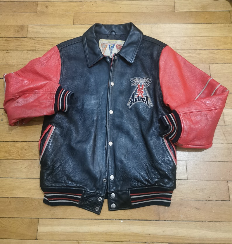 May include: A black leather varsity jacket with red leather sleeves. The jacket has a snap closure, two pockets, and a ribbed collar, cuffs, and hem. The jacket has a large "AVIREX" patch on the left chest with a USA flag and an eagle design.