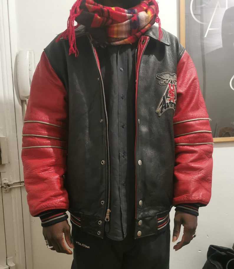 May include: A black leather varsity jacket with red sleeves and a white and silver stripe detail. The jacket has a zipper closure and a patch on the left side that says "USA Aviator".