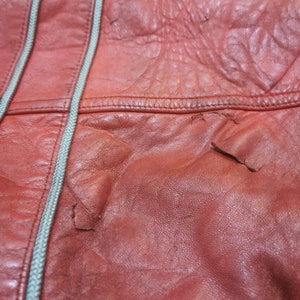 May include: Close-up of a red leather bag with a white zipper. The leather has a textured surface and shows signs of wear.